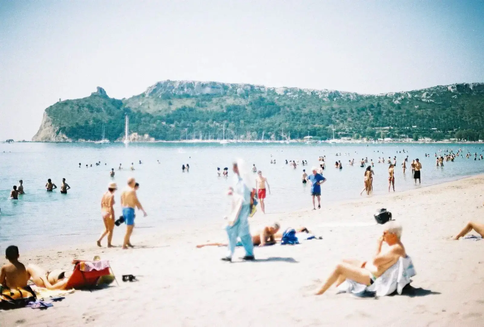 A busy beach on a bright day: swimmers in a pale blue bay, sunbathers on the sand and a green headland across the water